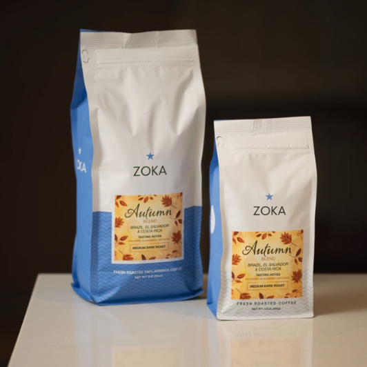 Two bags of Zoka Autumn Blend Coffee, one larger blue and white bag and one smaller white bag, standing on a white reflective surface against a dark background.